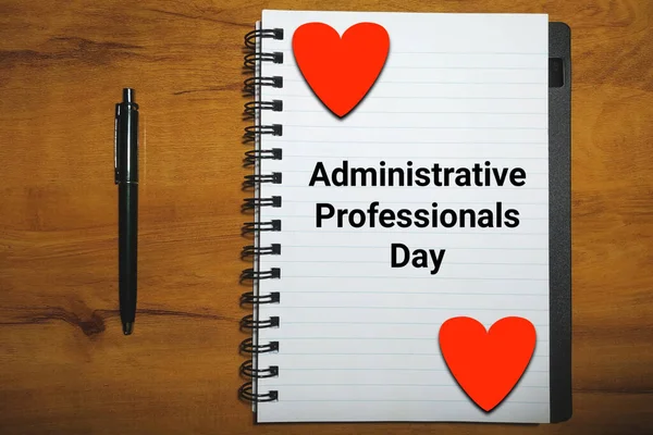 Administrative Professionals Day on notebook with pen and hearts on wood background