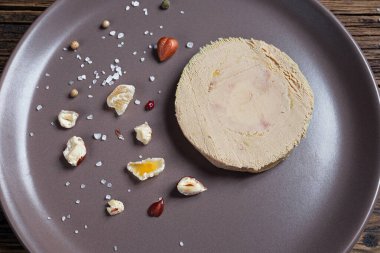 French foie gras served with nuts, pepper, salt, and dried fruits on a brown plate, top view close-up
