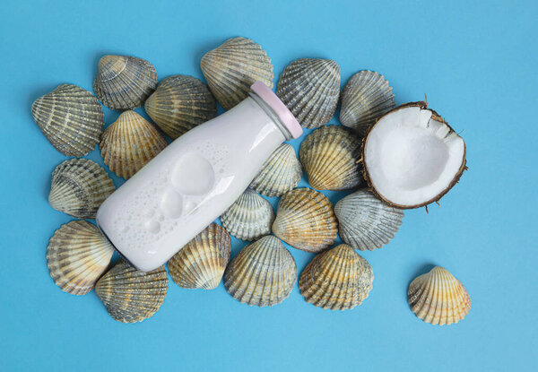 A bottle of vegan coconut milk and a piece of coconut lies on seashells on a blue background, alternative milk concept, top view