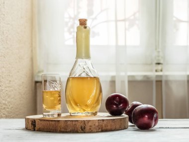 Balkan erik brendisi - rakija veya rakia slivovica şişede, sljivovica 'lı bir şarap kadehi ve pencerenin yanındaki açık arkaplanda taze erikler.