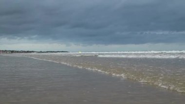 Hafif dalgaların kıyıya vurduğu sakin plaj sahnesi, yukarıda dramatik bulutlu bir gökyüzü, rahatlama ve tefekkür için huzurlu bir ortam çağrıştırıyor.