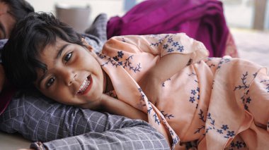 Cute Indian baby sleeping peacefully in bed with her mother at home. 5 year old Innocent daughter hug and laugh while lying on the striped bed. Little baby girl relaxing