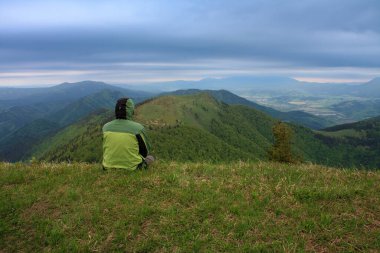 Otların üzerinde oturan yürüyüşçü, arka plandaki Osnica dağı, Mala Fatra, Slovakya bahar bulutlu sabahı