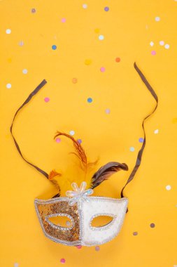 Zarif tüylerle süslenmiş altın ve beyaz bir maske, canlı sarı bir arka plana karşı duruyor.
