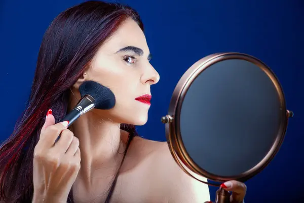 Woman applying makeup reflected in a mirror against a blue background