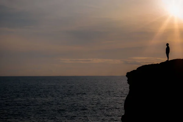 Silhouette of man stands on the edge of the abyss and looks at the sea on big stone at sunset in mountains.
