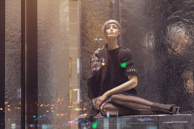 Lüks bir mağazanın vitrininde oturan kadın manken ve şehrin gece ışıkları.