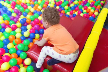 Kıvırcık saçlı bir bebek, çocukların oyun odasındaki çok renkli bir havuzda oynar. İnsanlar