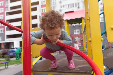 Kıvırcık çocuk çok katlı bir binanın yanındaki oyun alanında oynuyor. İnsanlar