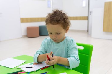 Kıvırcık çocuk masada oturan kalemlerle resim çizer. İnsanlar