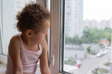 Kıvırcık çocuk pencerenin kenarında otururken pencereden dışarı bakar. İnsanlar