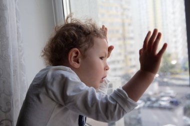 Kıvırcık bir çocuk duygusal olarak pencereden dışarı bakar, pencere pervazında oturur. İnsanlar