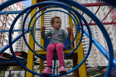 Kıvırcık bir çocuk çok katlı bir binanın arka planında oyun oynuyor. İnsanlar