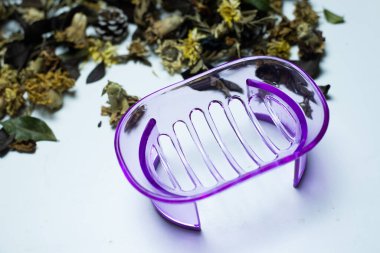 A clear purple soap box, empty, set against a white, blurry background, with dried flowers surrounding it