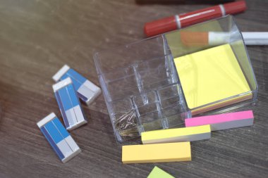 Ofis malzemeleri koyu kahverengi çalışma masasına yayılmış. Çeşitli küçük not kağıtları, kalemler, silgiler, belge kabloları var..