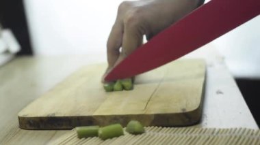Yeşil sebzeleri kırmızı bıçakla kesen ellerin videosu. Aşağıda ahşap bir kesme tahtası var.