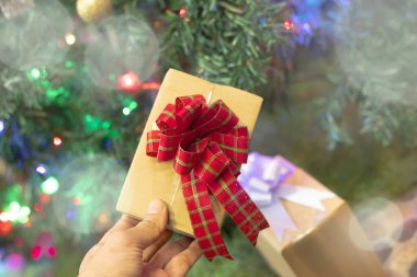 Bir insanın eli basit kahverengi bir kağıda sarılı küçük bir hediye tutar ve şenlikli kırmızı ve yeşil ekose bir kurdeleyle bağlanır. Arka plan güzel bulanık ve parıldayan bir Noel ağacı, renkli ışıklar ve süslerle süslenmiş, yumuşak bir bokeh yaratıyor.
