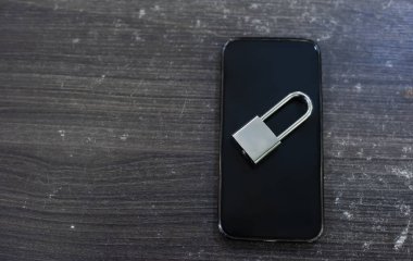 Ekranında gümüş metal bir asma kilit olan modern bir siyah akıllı telefonun yukarıdan çekilmiş yakın çekim görüntüsü. Nesneler, temiz bir arkaplan görevi gören koyu, ahşap bir yüzeye yerleştiriliyor. Kompozisyon basit ve kavramsal, t ile.