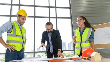 İnşaat mühendisi ve mimar yelek ve miğfer içinde inşaat müdürüyle planlarını tartışıyorlar, inşaat alanında duruyorlar.