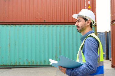 Foreman ya da Mühendis beyaz başlık takıyor ve elinde evrak kağıtları olan yansıtma gömleği giyiyor ve arka plandaki konteynırlarla envanter ve iş detaylarını kontrol etmeye çalışıyor. Lojistik Ulaşım Konsepti.