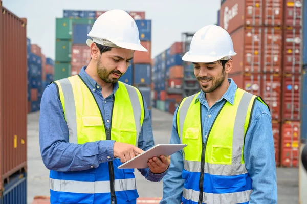 Container operator wearing white helmet and reflection shirt and holding tablet doing routine inspection while working in container yards. Logistics and shipping concept