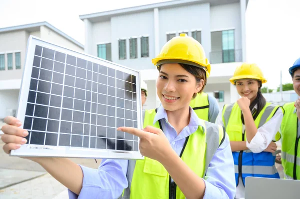 electrician woman in safety vest with helmet showing solar cell panel with the team in background, standing in the under-construction building. Green energy electricity concept. concept of sustainable resources