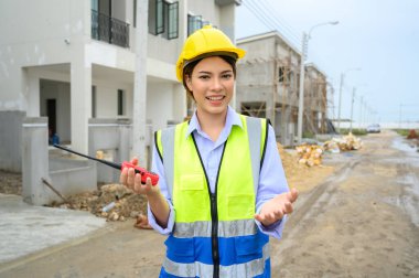 Young construction engineer woman in safety vest with yellow helmet holding radio, standing on building construction site. Smiling talking to the camera