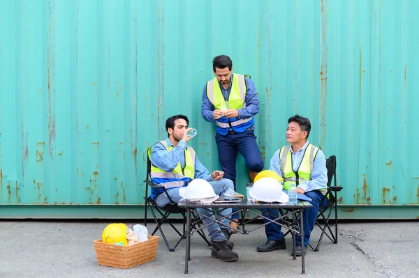Group of logistic staff workers talk and rest sitting in the shipping yard container during break time. Cargo ship import export factory logistic transport and global business concept.