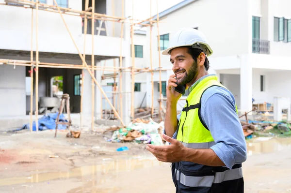 Young attractive construction smiling bearded using smartphone calling to client, worker in vest with white helmet standing on construction site