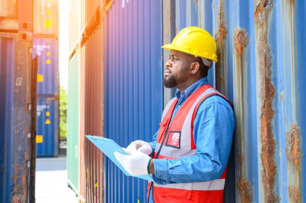 African American logistic workers wear yellow helmets and reflection shirts, hold document paper and look to check inventory or job details at the shipping container yard. Transportation import and export logistic industry concept