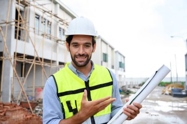 Young constructor engineer man smiling holding blueprint while talking about building construction, looking camera on under-construction building site. Happy Constructor bearded in vest with white helmet