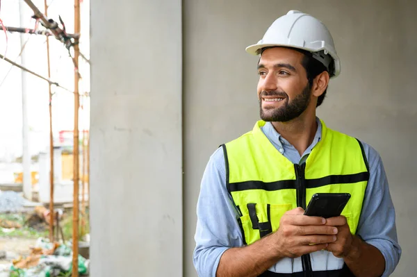 portrait of young engineer in vest with white helmet standing on construction site, smiling and holding smartphone for worker, internet, social media