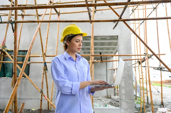 Young attractive construction woman smiling with yellow helmet working with laptop, standing on building construction site. Home building project
