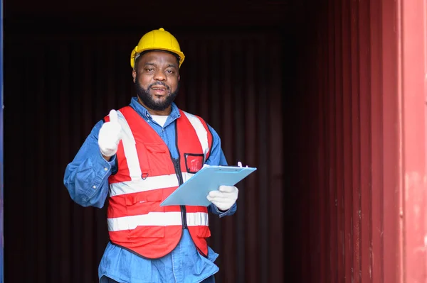 African American logistic workers wear yellow helmets and reflection shirts, hold document paper and look to check inventory or job details at the shipping container yard. Transportation import and export logistic industry concept