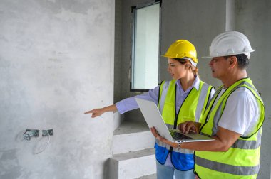 Yelekli, kasklı, dizüstü bilgisayarlı genç ve çekici bir inşaat işçisi. İnşaat alanında duruyor. Ev yapımı projesi. Mühendis ustabaşı iş yerinden bir iş arkadaşıyla konuşuyor.