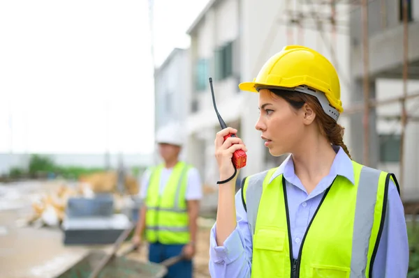 Young attractive construction woman in safety vest with yellow helmet working with radio, standing on building construction site. Home building project
