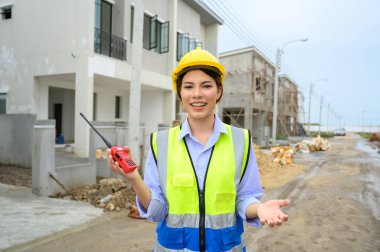 Young construction engineer woman in safety vest with yellow helmet holding radio, standing on building construction site. Smiling talking to the camera