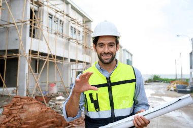 Young constructor engineer man smiling holding blueprint while talking about building construction, looking camera on under-construction building site. Happy Constructor bearded in vest with white helmet