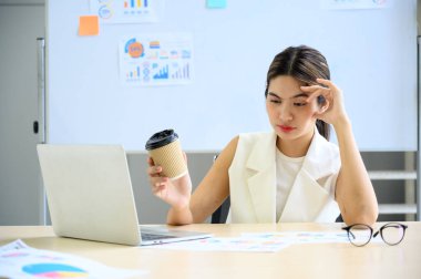 Kahve fincanı tutan yorgun ve yorgun genç iş kadınları ofis masasında baş ağrıları çekiyorlar. Kendini yorgun hissederken çalışan Asyalı kadın.