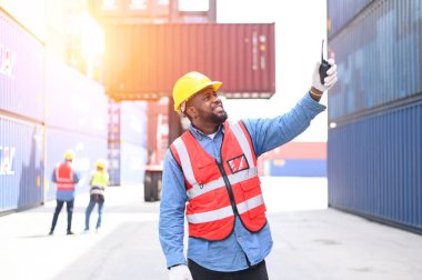 Sarı kask ve güvenlik yelekleri takan Afro-Amerikan konteynır operatörleri konteynır sahalarındaki telsiz çalışanları aracılığıyla kontrol ediyorlar. Ulaştırma ithalat ve ihracat lojistik sanayi kavramı.