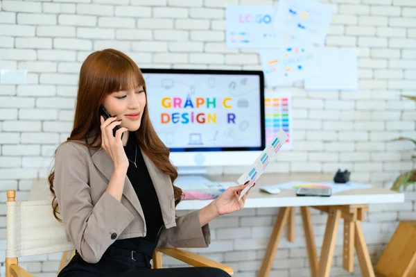 Asian female graphic designer talking on mobile phone with colleague or customer while working in office