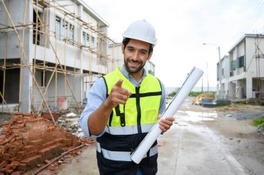 Young constructor engineer man smiling holding blueprint while talking about building construction, looking camera on under-construction building site. Happy Constructor bearded in vest with white helmet
