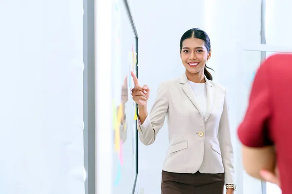 Young Asian businesswoman manager pointing finger at paperwork, pie chart diagram document on glass board in the office. Looking at the camera