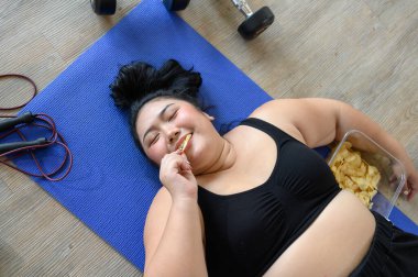 Egzersiz sırasında yoga minderinde uzanmış patates cipsi yiyen tombul Asyalı kadının en iyi görüntüsü. Spor elbiseli şişman genç kadınlar sağlıksız olmaktan memnunlar ve ellerinde bir kase gevrek atıştırmalık ve fast food yiyorlar.