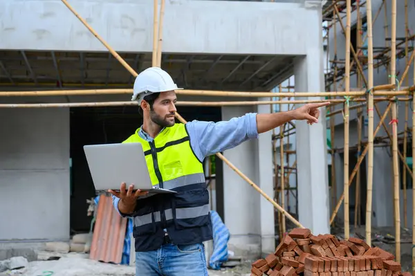 Young attractive construction smiling bearded in vest with white helmet working with laptop, standing on building construction site. Home building project