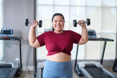 Genç, pozitif, gülümseyen, neşeli, kilolu kadın spor salonunda dambıllarıyla antrenman yapıyor. Spor kıyafetli şişman kadın spor yapıyor. Kilo verme egzersizi, sağlıklı yaşam tarzı konsepti