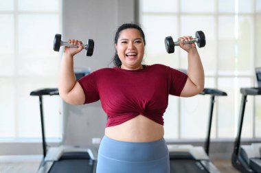Genç, pozitif, gülümseyen, neşeli, kilolu kadın spor salonunda dambıllarıyla antrenman yapıyor. Spor kıyafetli şişman kadın spor yapıyor. Kilo verme egzersizi, sağlıklı yaşam tarzı konsepti