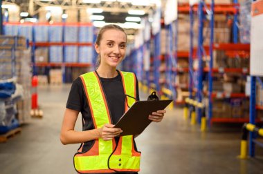 Fabrika işçisi kadın elinde pano tutarken işyeri deposundaki hisse senedi siparişleri detaylarını ve malzeme malzemelerini kontrol ediyor.