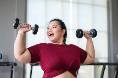 Genç, pozitif, gülümseyen, neşeli, kilolu kadın spor salonunda dambıllarıyla antrenman yapıyor. Spor kıyafetli şişman kadın spor yapıyor. Kilo verme egzersizi, sağlıklı yaşam tarzı konsepti