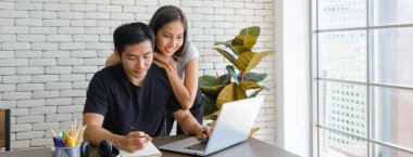 Genç Asyalı çiftler evde çalışırken günlük kıyafetler giyerler. Rahat olun güzel beyaz odada dizüstü bilgisayarlarla çalışan çiftler